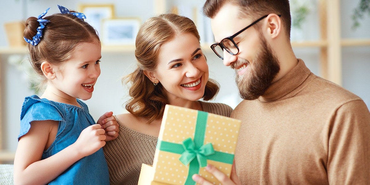 Family of 3, Mother, Father and daughter stood together, smiling, opening a Father's Day gift from a box with a ribbon.